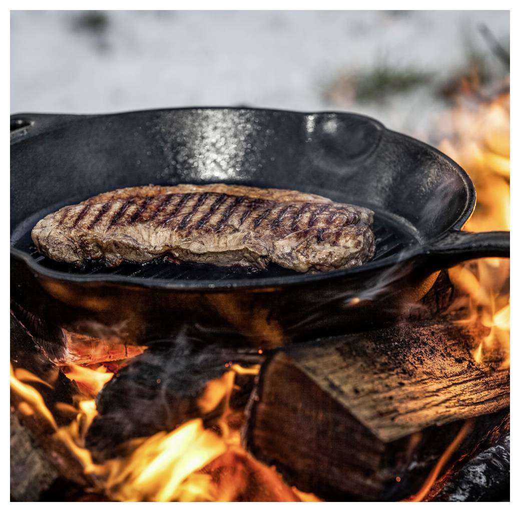 Steak se smažící v litinové pánvi nad otevřeným krbem z tvrdého dřeva, s plameny a kouřem pod pánví.