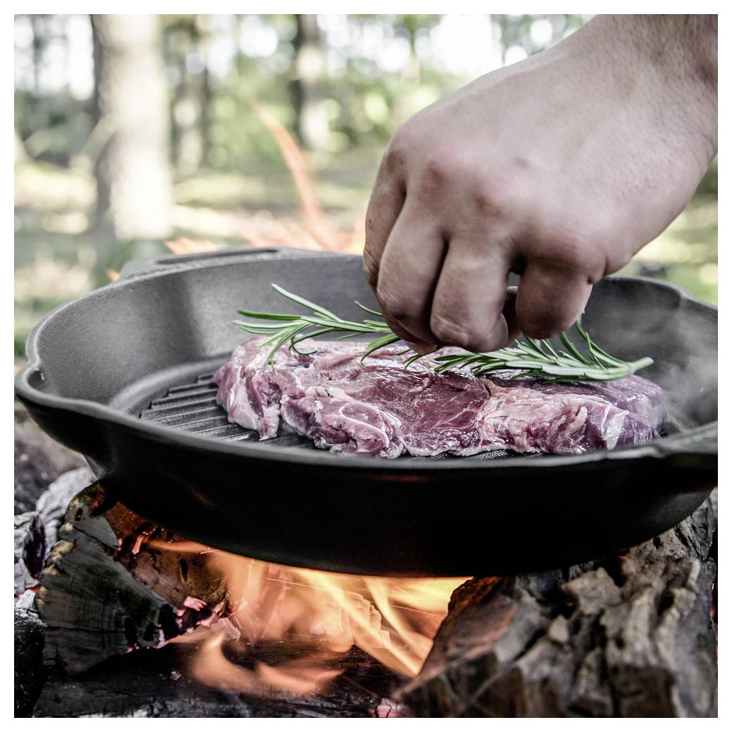 Ruka ochucuje surový steak rozmarýnem v litinové pánvi nad otevřeným táborákem.