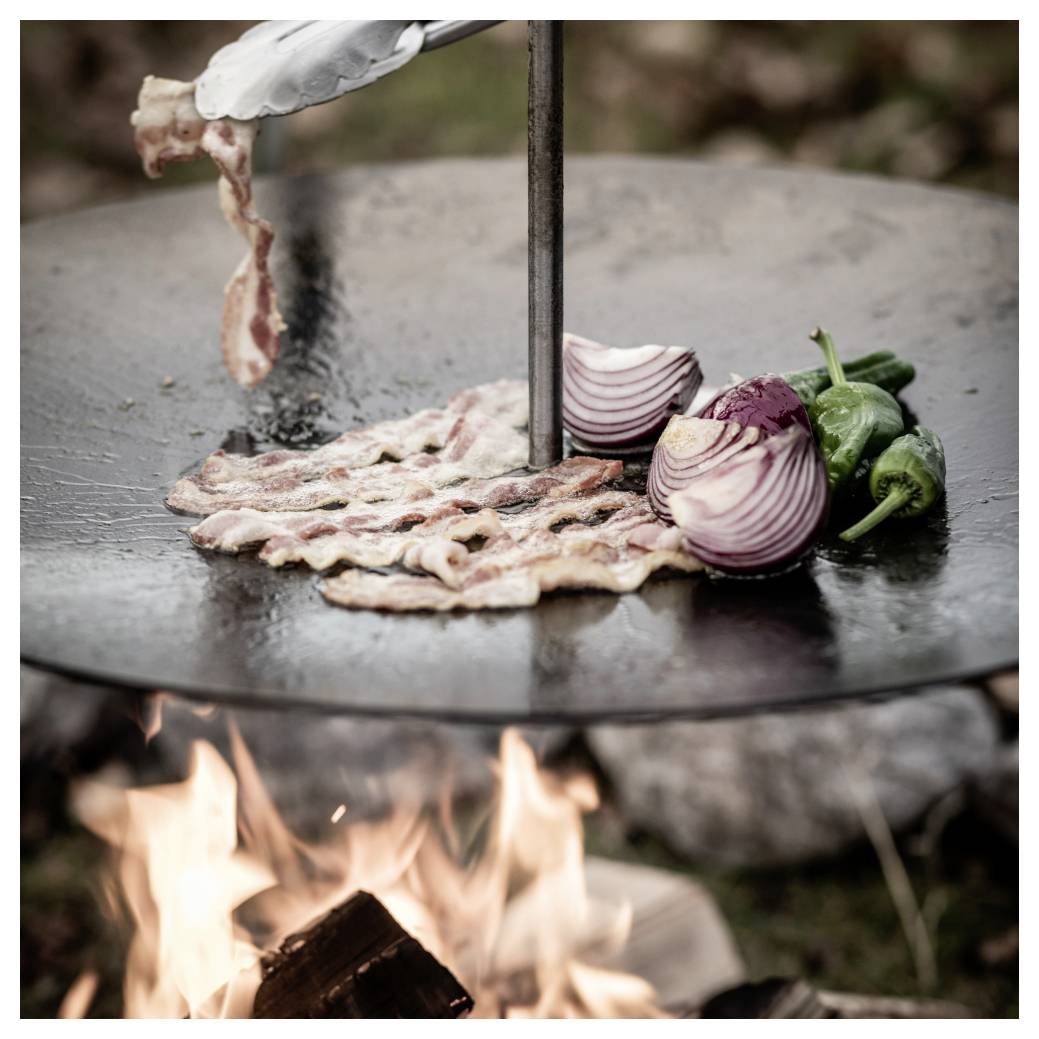 Slanina vaří se na horké venkovní grilovací desce nad otevřeným ohněm, vedle ní je krájená červená cibule a zelené papriky.