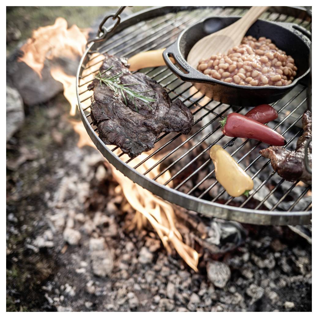 Steak, fazole a paprika se vaří na grilu nad otevřeným táborákem, pod mřížkou jsou viditelné plameny.