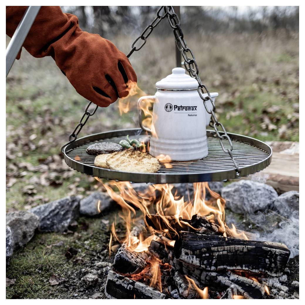 Rukavicová ruka vaří jídlo a zahřívá petrolejový hrnec na visícím grilu nad otevřeným táborovým ohněm venku.