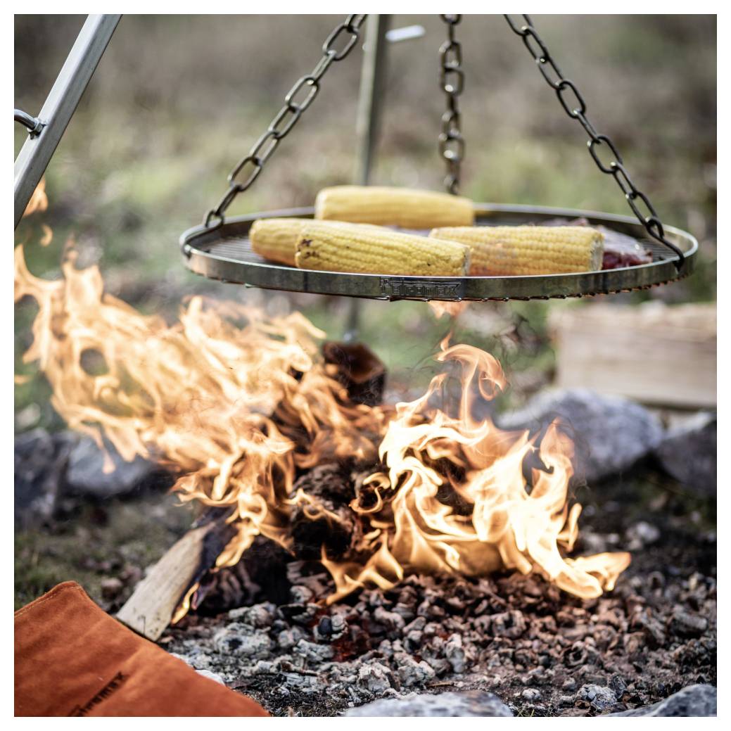 Kukuřice na trní se vaří na visícím grilu nad otevřeným táborovým ohněm s plameny a žhavými uhlíky.