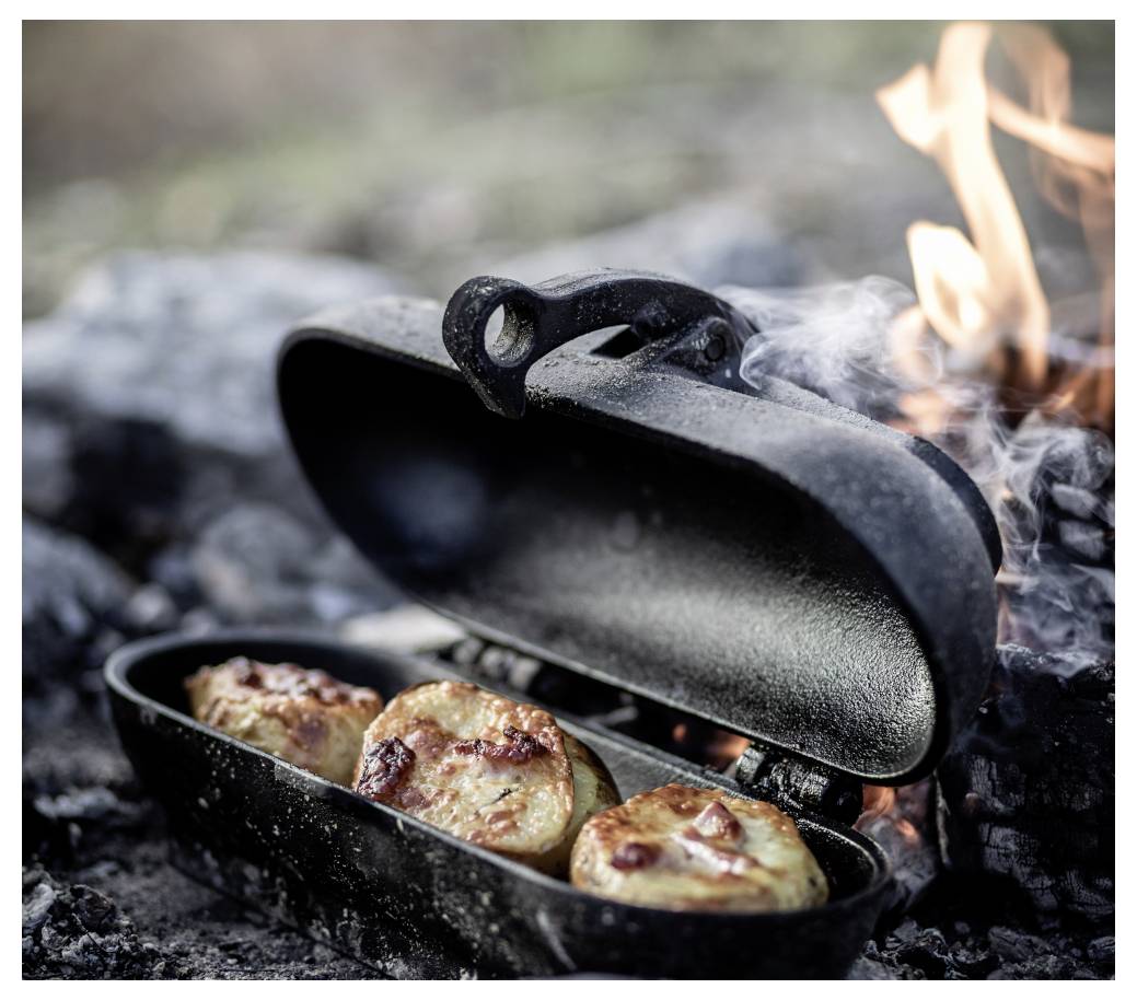 Litinový dutchoven na otevřeném ohni s otevřeným víkem, obsahující hnědé péčené knedlíky nebo těstoviny, s plamenům a kouřem stoupajícím nahoru.