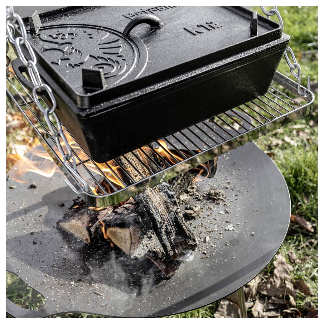 Litinový Dutch oven s reliéfním víkem visí nad dřevěným ohněm na kulatém venkovním ohništi, vaří se nad plameny a kouřem.
