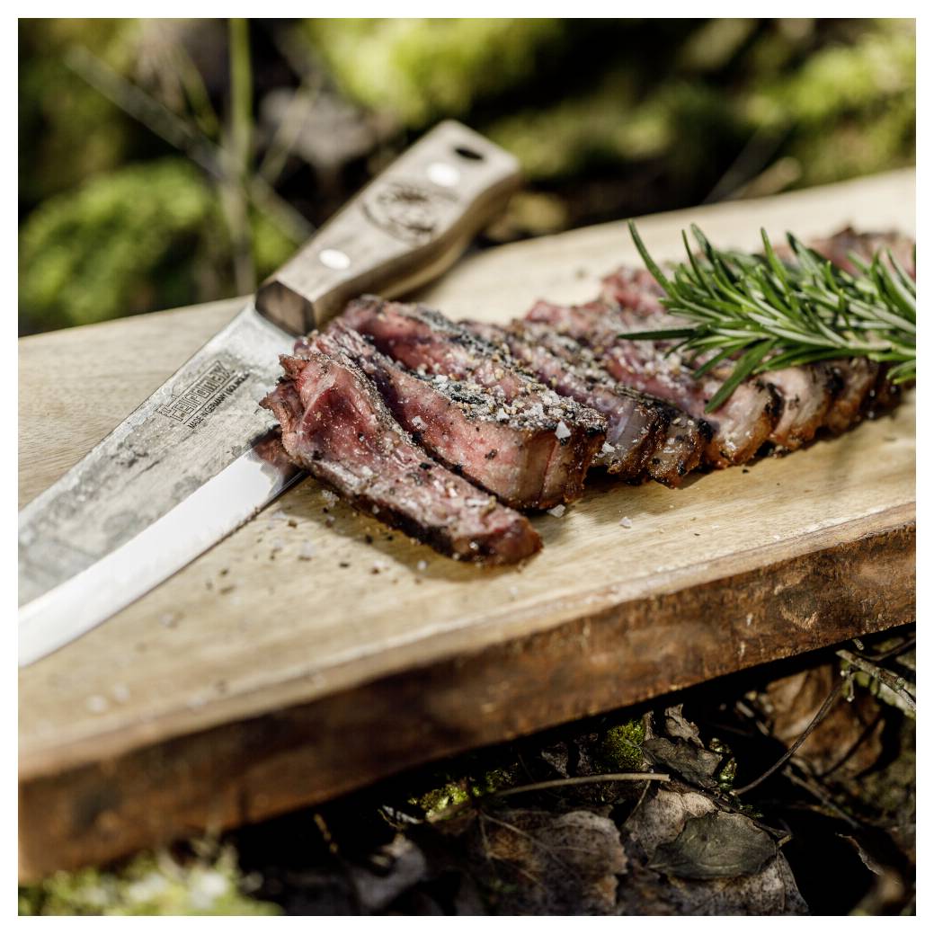 Nakrájené grilované steak na dřevěné kuchyňské desce s nožem a garnáží z rozmarýnu venku.