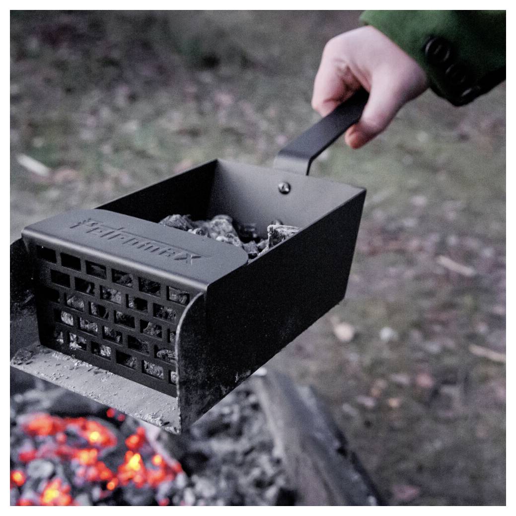 Ruka držící kovový komín na uhlí vyplněný horkými uhlíky nad ohněm, připraven k zapálení grilu nebo ohniště.