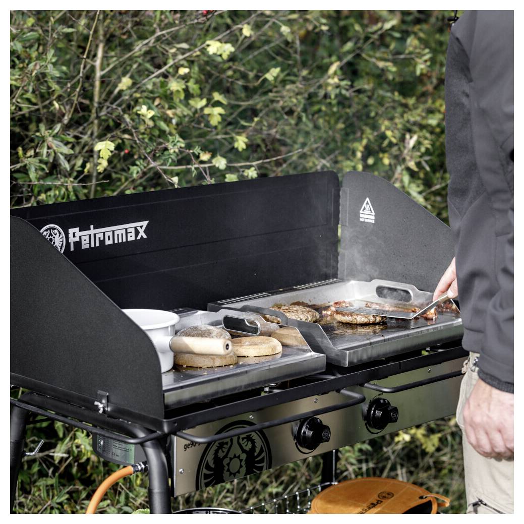 Osoba griluje hamburgery a buláčky na plynovém grilu Petromax venku, viditelná je stěrka a postranní tácky.