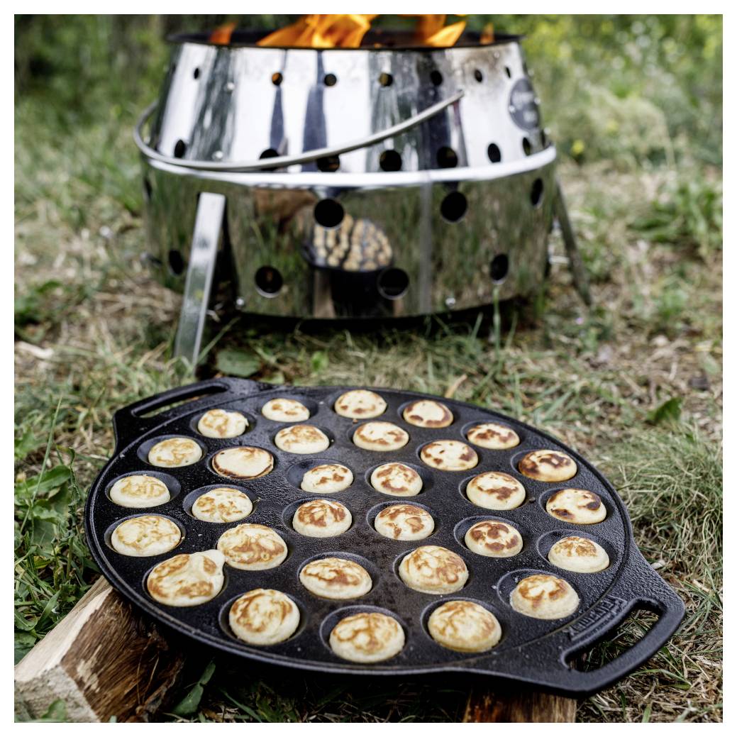 Scéna venkovního vaření s litinovou plotnou se malými palačinkami nebo blini na trávě vedle rozžehnuté kovové ohniště.