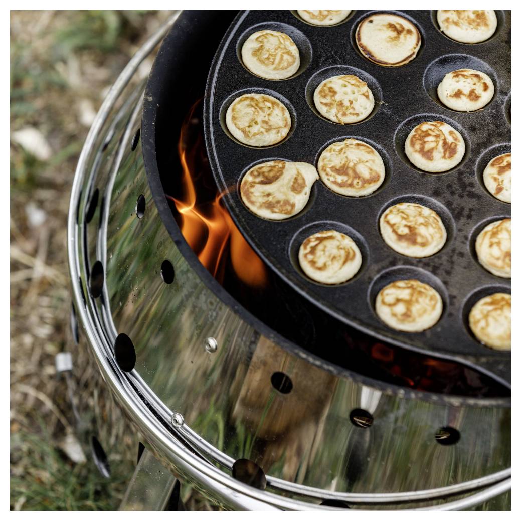Těsto na mini palačinky vaří se v kulaté litinové pánvi nad otevřeným ohněm na grilu.
