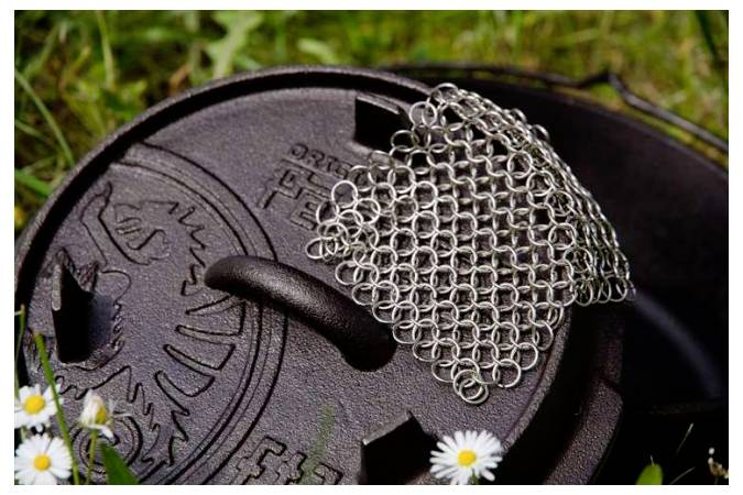 Detailní snímek víka litinového hrnce nebo holandské trouby s drátěnkou na čištění položenou navrchu, umístěný venku v trávě s malými bílými květy.