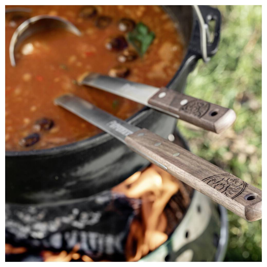 Dvě nože s dřevěnými rukojetí spočívají na hrnci vařícího se chilli na venkovním kempinkovém ohništi.