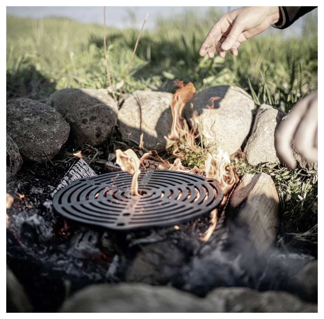 Ruka dosahující k malému táboráku hořícímu pod kulatou kovovou mřížkou grilu mezi kameny venku.