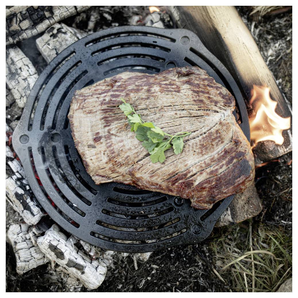 Grilovaný steak zapékající se na kulatém litinové mřížce nad otevřeným táborákem, ozdobený malou větvičkou byliny.