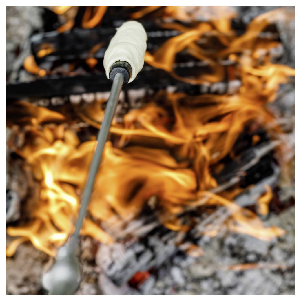 Marshmallow pražený na tyči nad otevřeným kempovým ohněm s jasným plamenem a žhavými uhlíky.
