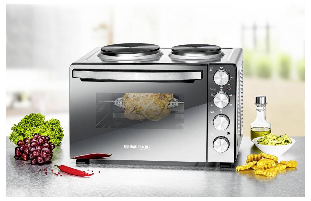 Stůl na desku s pečeným kuřetem viditelným uvnitř, zobrazený s kuchyňskými ingrediencemi a pomůckami jako jsou papriky, hrozny, olej a krájené brambory.