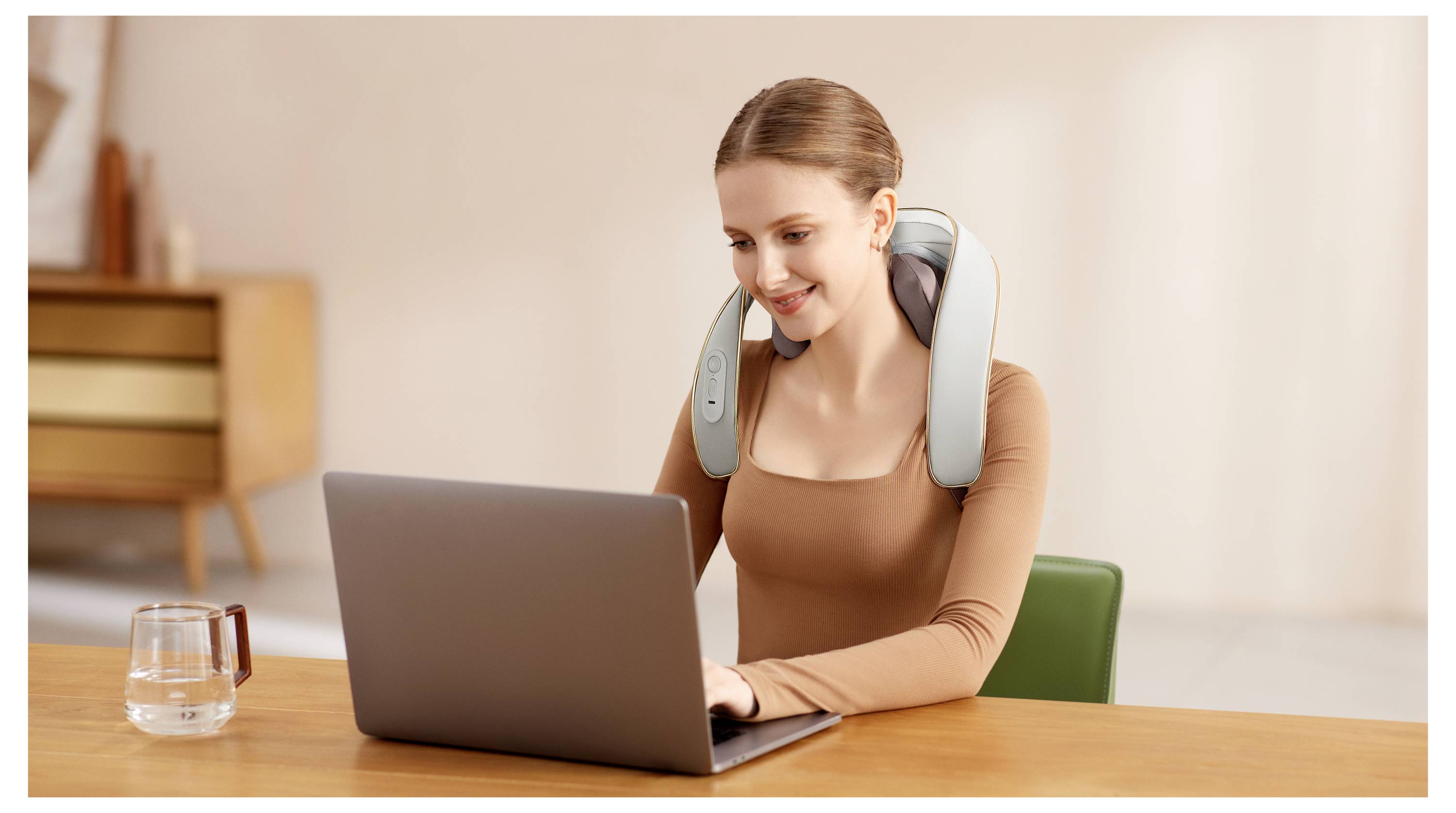 Žena s masážním přístroje na krku používá laptop u stolu v domácím prostředí s sklenicí vody v blízkosti.