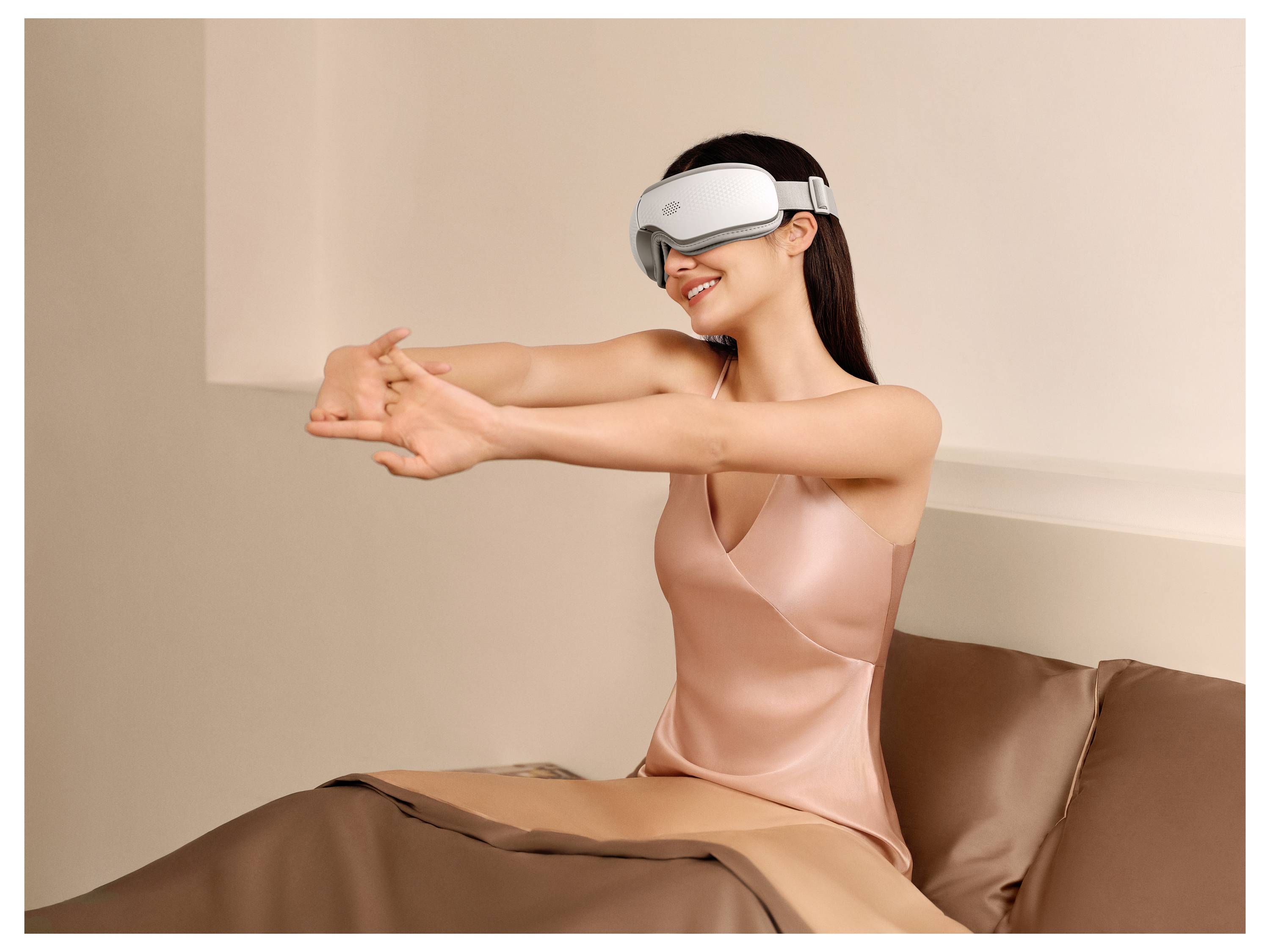 Žena v headsetu virtuální reality sedí na posteli a natahuje ruce dopředu, přičemž se usměje v jemně osvětlené ložnici.