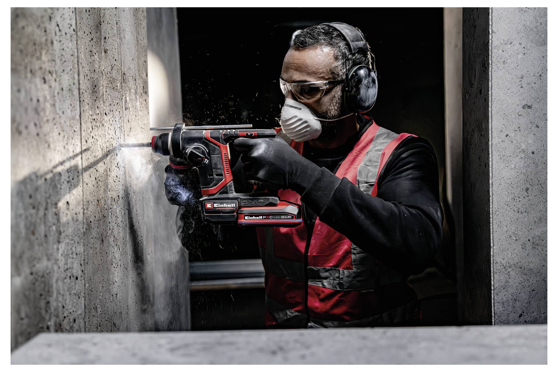 Stavbyň v ochranném vybavení vrtá do betonové stěny červeným elektrickým nástrojem v úzké pracovní oblasti.