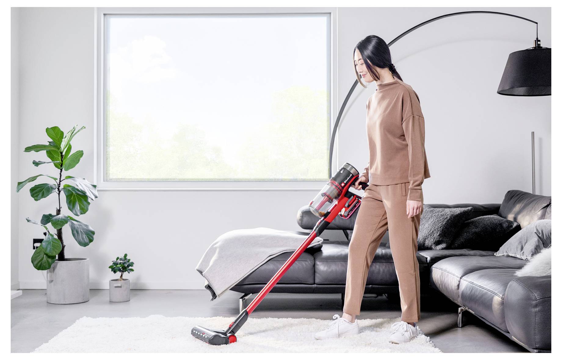 Žena vysavačem čistí bílý koberec v jasné obývací místnosti vedle černé pohovky a stojící lampy.