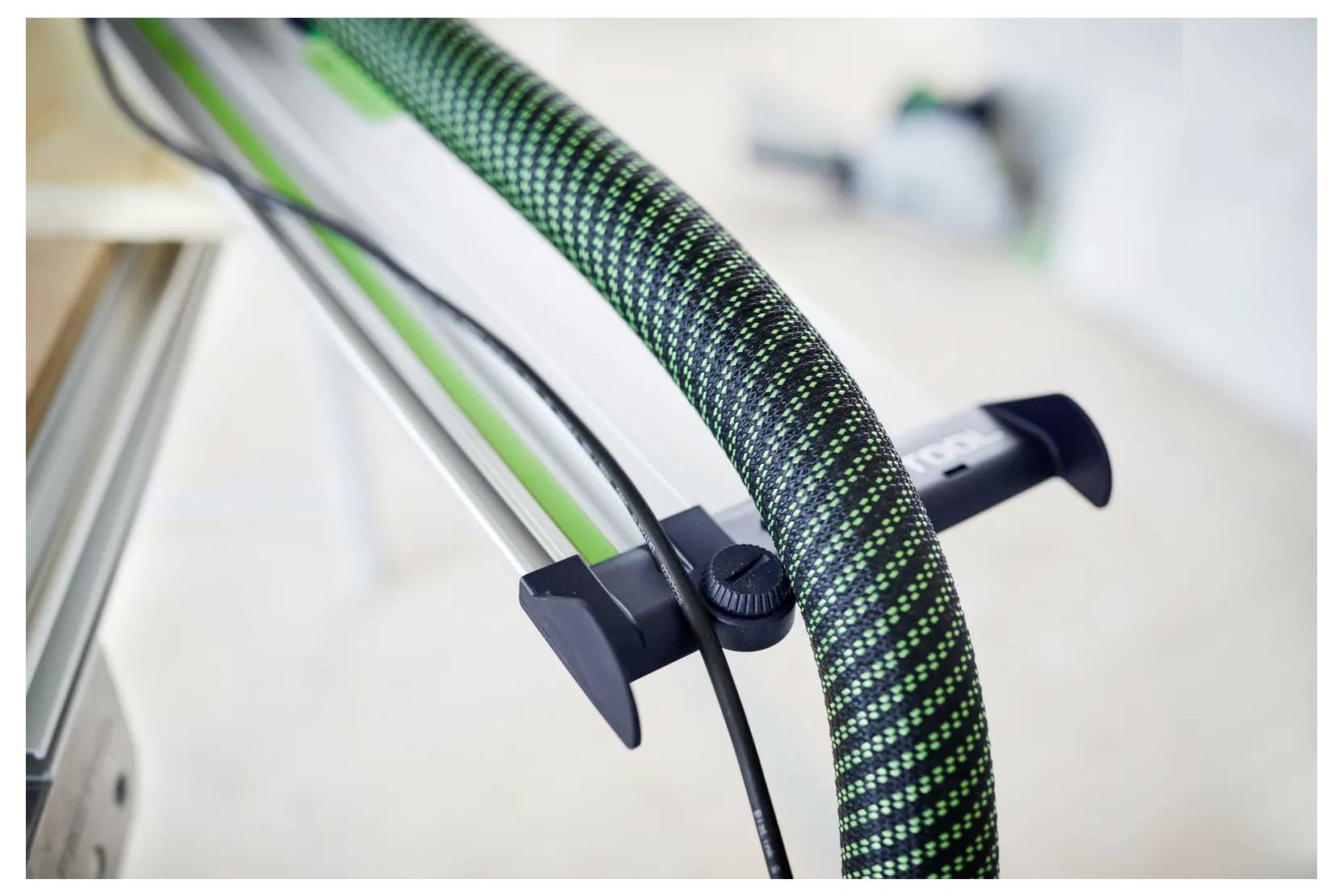 Detailní záběr na zeleno-černě pletené hadice nebo kabel připevněný na kovový rám pomocí klipu, přičemž je v ohnisku černá nastavovací přilba.