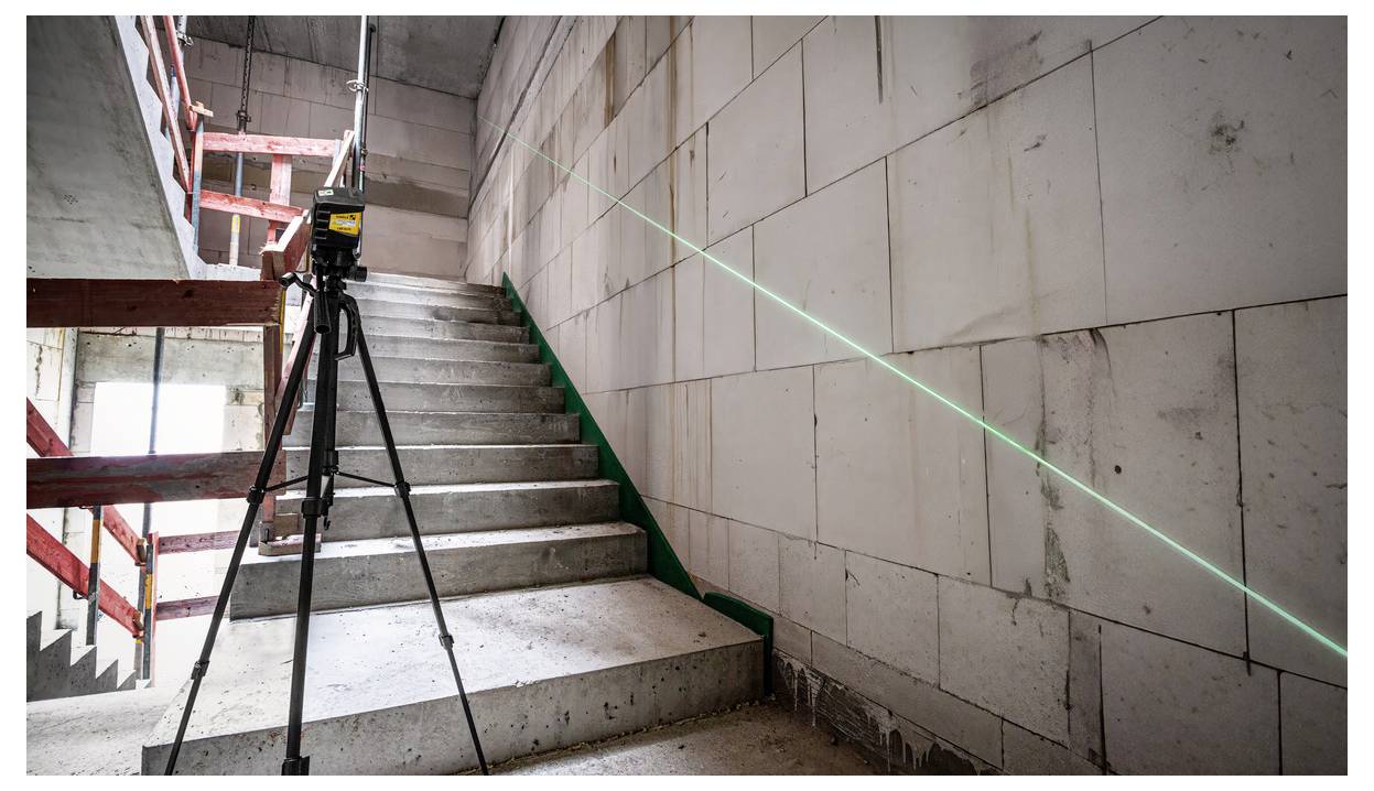 Nivelační laser na stativu promítající zelenou linii podél nedokončeného betonového schodiště uvnitř staveniště.