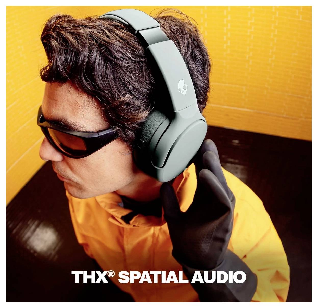 Osoba v oranžové bundě nosící sluneční brýle a násluchátka přes uši, se textem „THX SPATIAL AUDIO" v dolní části.