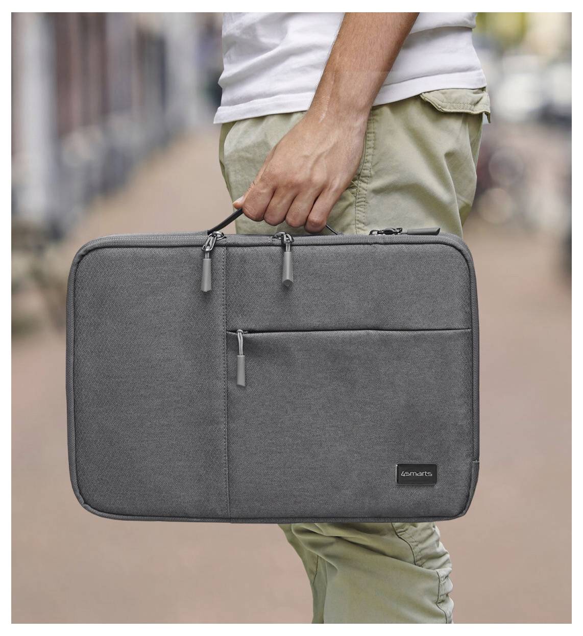 Detailní snímek osoby držící šedý laptop brašnu se zipem za rukojeť v exteriéru.