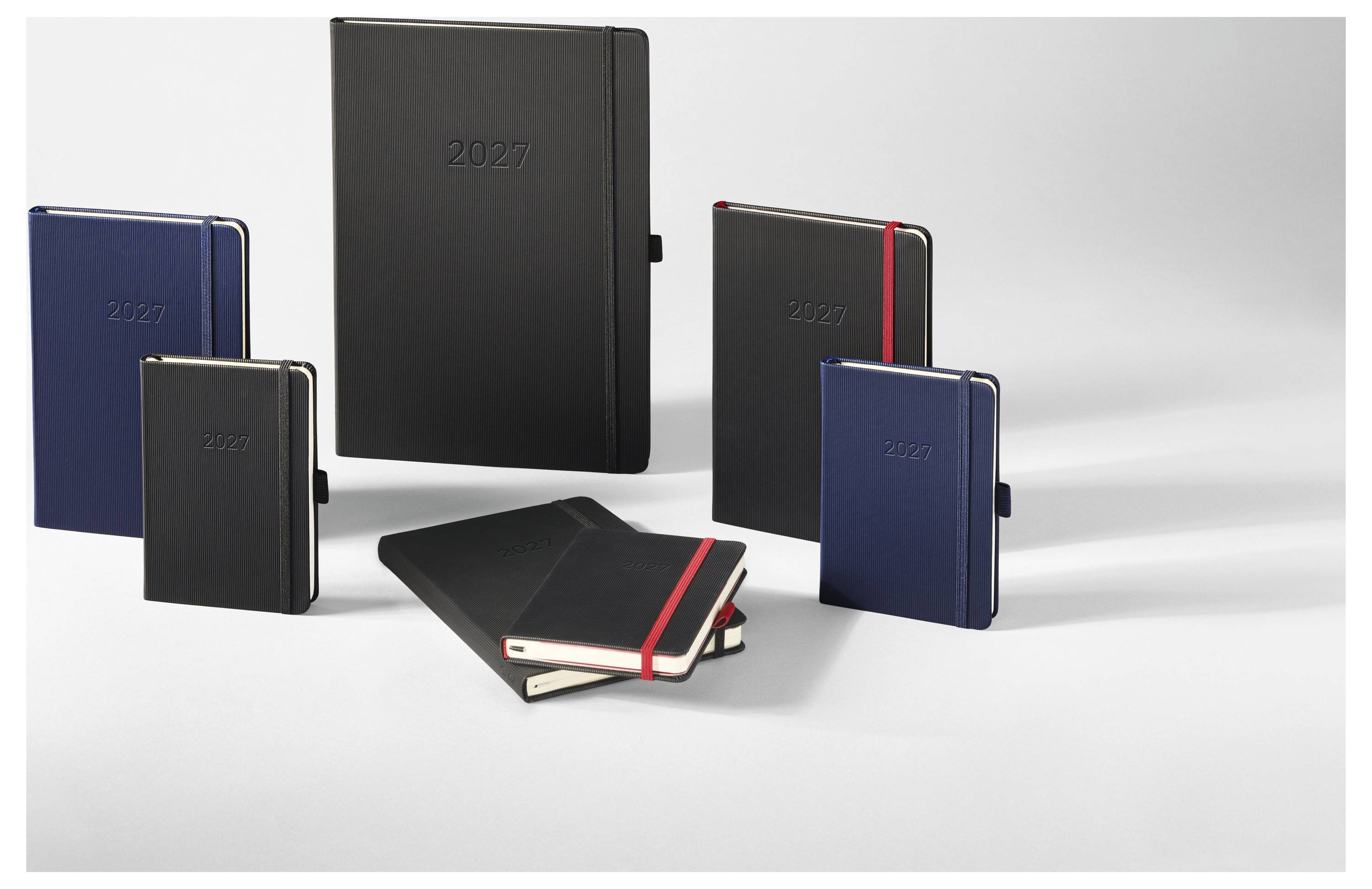 Sada 2027 plánovacích knih a notebooků v modré a černé barvě, včetně pevných desek a jednoho otevřeného notebooku, uspořádaných na bílém povrchu.