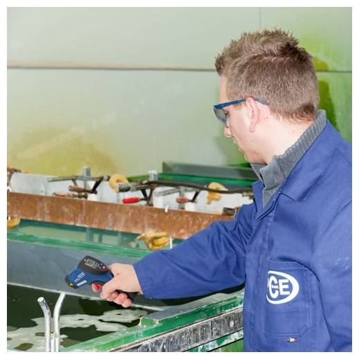 Technik v modré laboratorní halence používá přenosný tester nad vodní nádrží v průmyslovém nebo laboratorním prostředí.