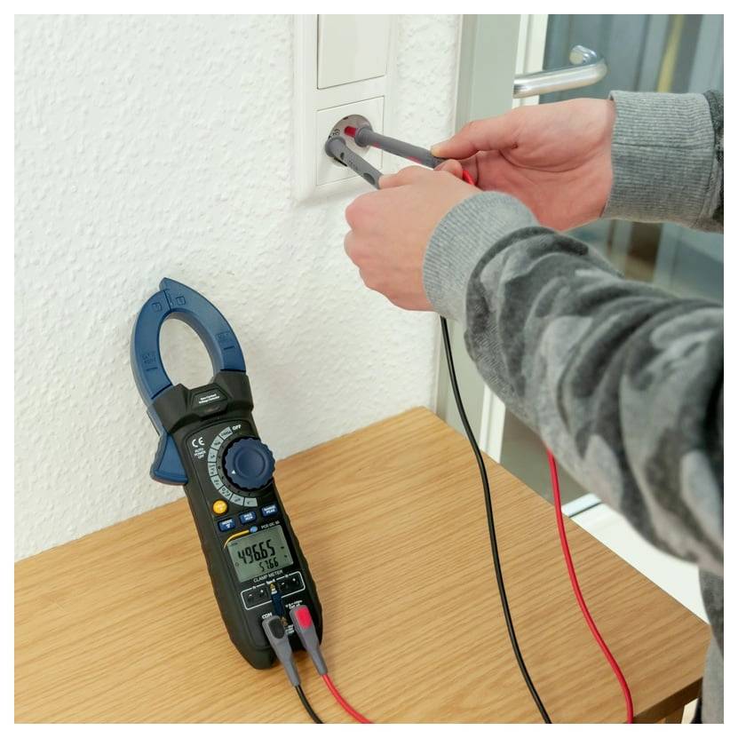 Osoba testující zásuvku na stěně s voltmetrem, zatímco kleštový multimetr leží na stole a zobrazuje elektrické nastavení pro řešení problémů.