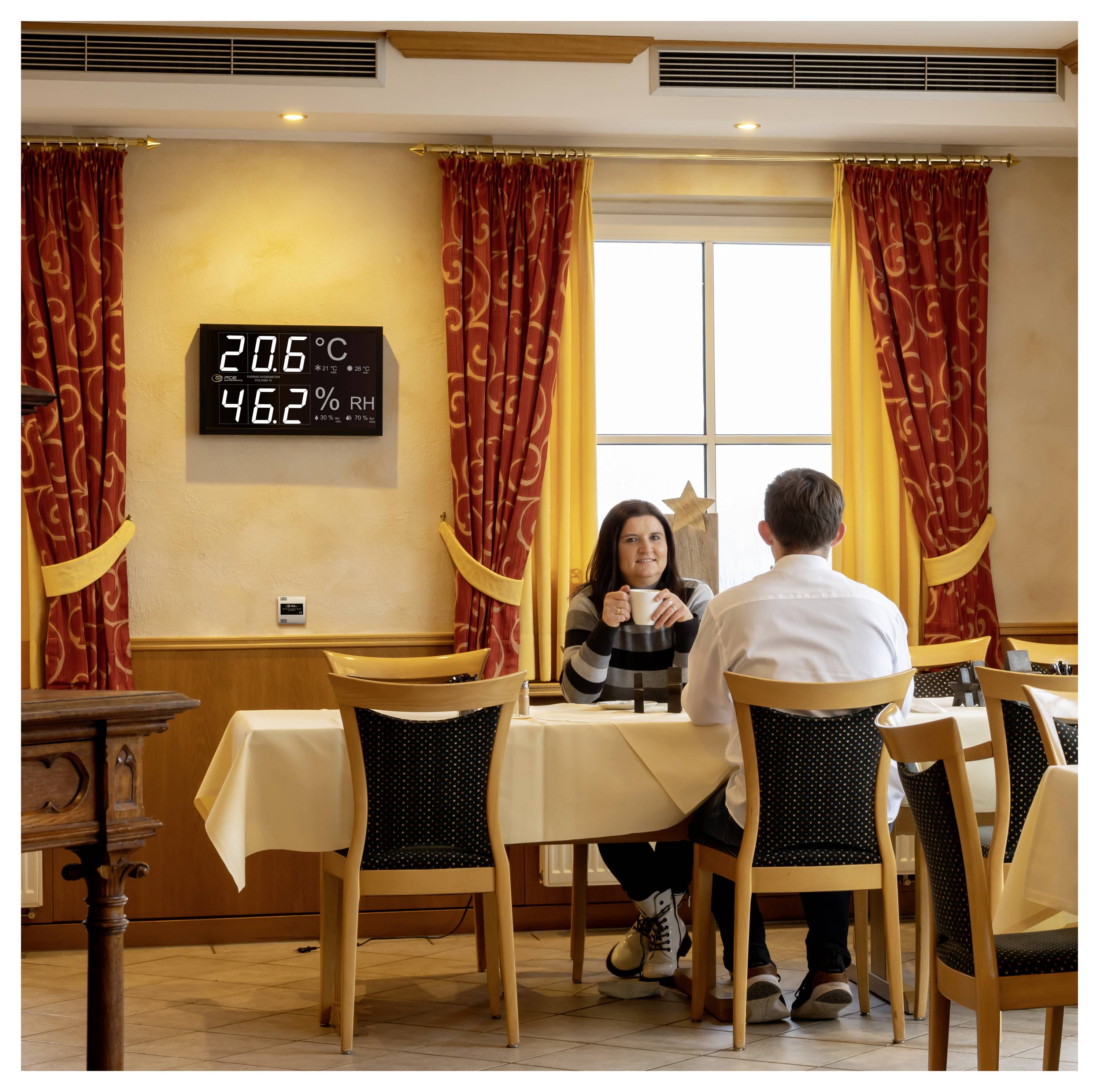 Dva lidé sedí proti sobě u stolku v restauraci u okna; nástěnný displej ukazuje „20,6°C" a „46,2% RH".