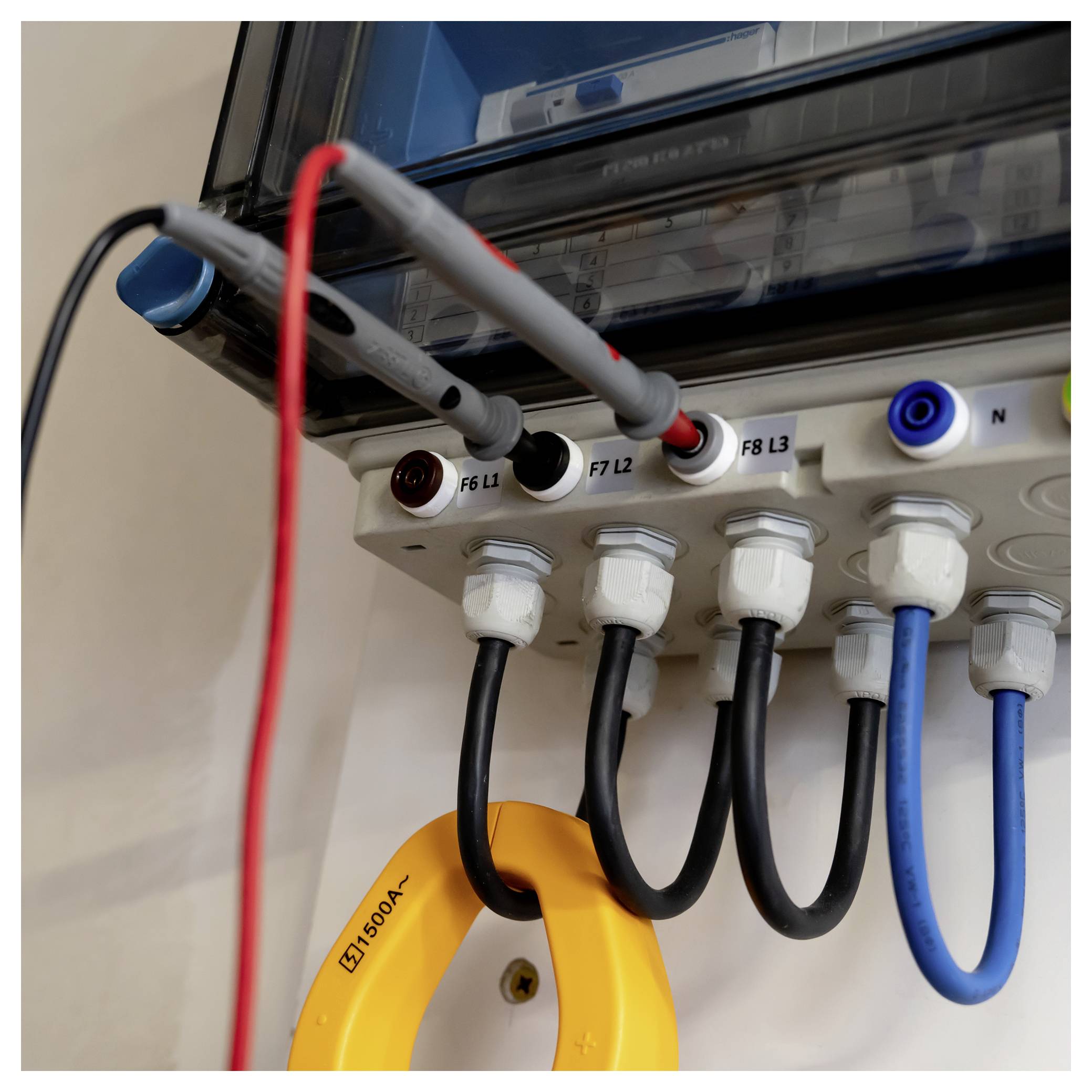 Detailní pohled na elektrický rozvodný panel s připojenými silnoproudými kabely, označenými svorkovnicemi a žlutými kleštěmi digitálního multimetru obešly kolem jednoho vodiče.