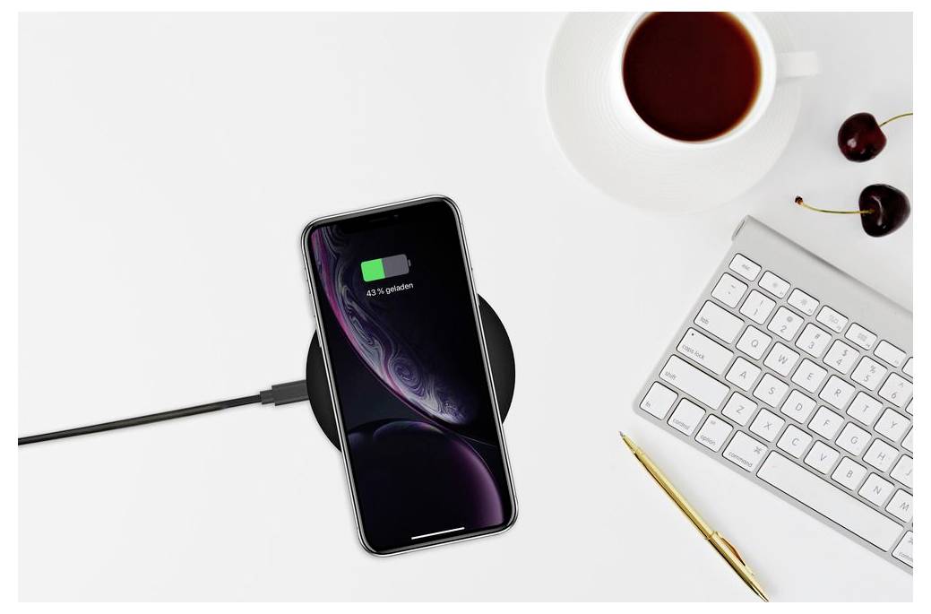 Smartphone připojený kabelem k nabíjení na bílém stole vedle klávesnice, pera, šálku kávy a třešní.