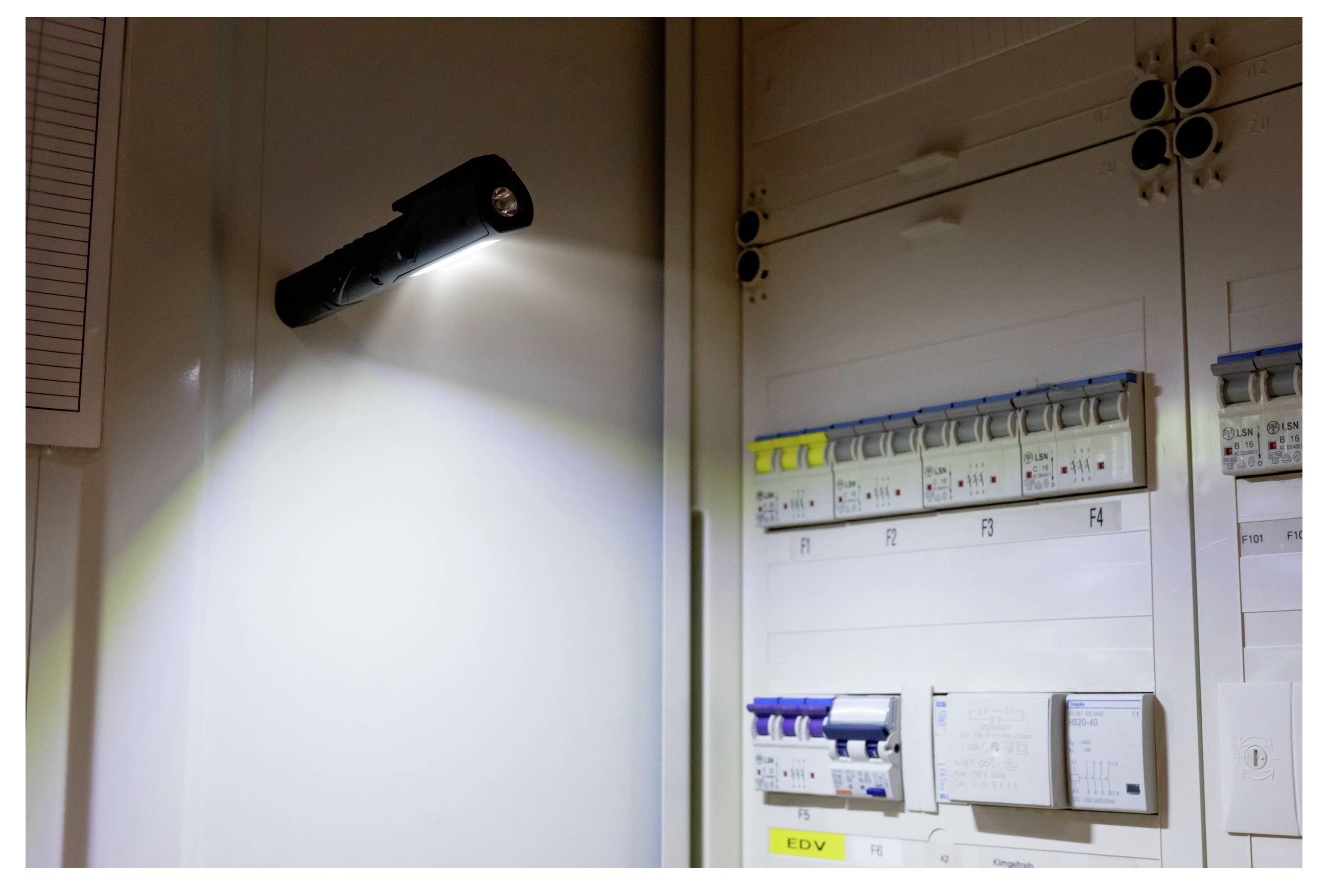 Nástěnné LED světlo svítící nad elektrickým rozvaděčem s označenými vypínači a řídicími moduly