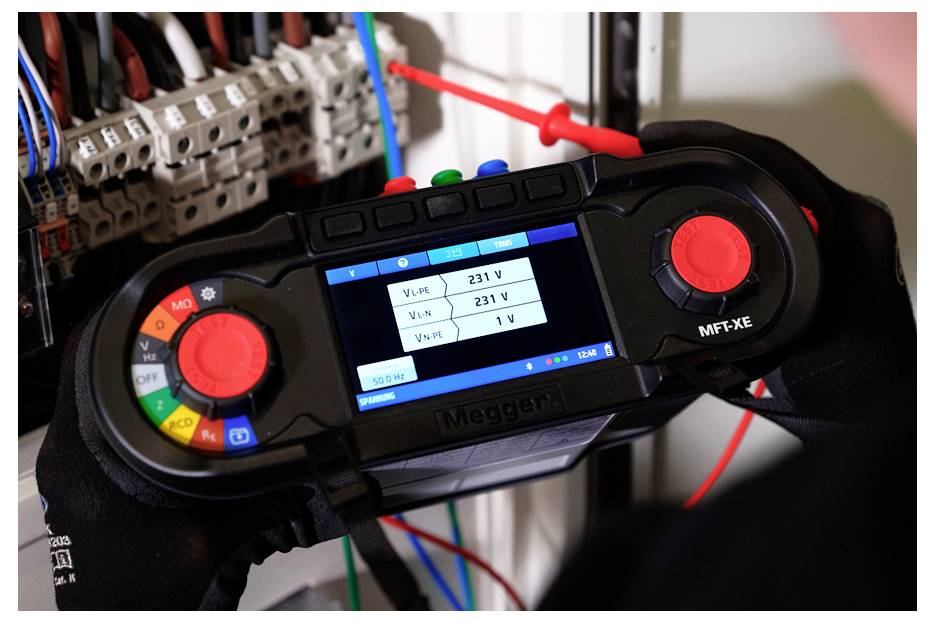 Rukavicemi oděné ruce drží Megger elektrický tester zobrazující údaje napětí v rozvaděči, s testovacími vodiči připojenými k jističům.