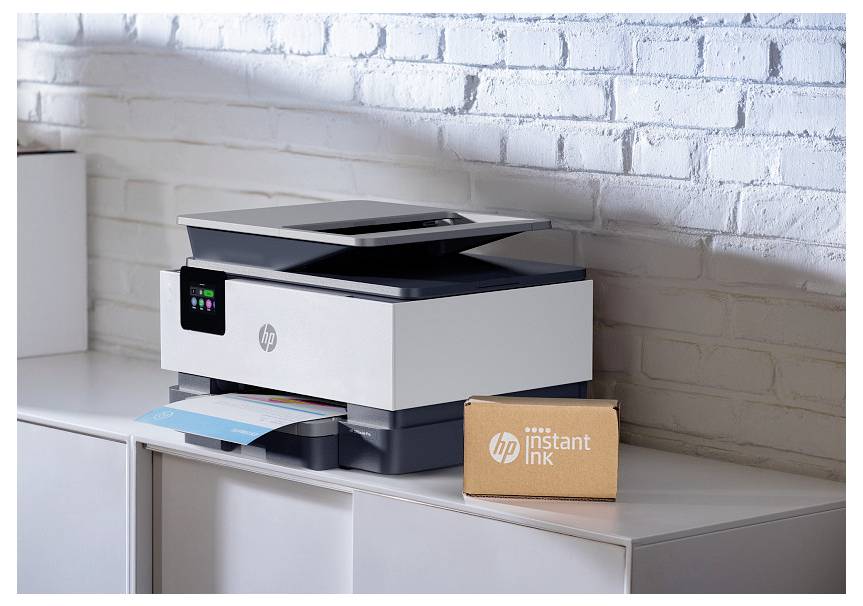 Tiskárna HP all-in-one na skříňce vedle krabice "HP Instant Ink", s částečně vytištěným dokumentem přitisknutým k bílé cihlové zdi.