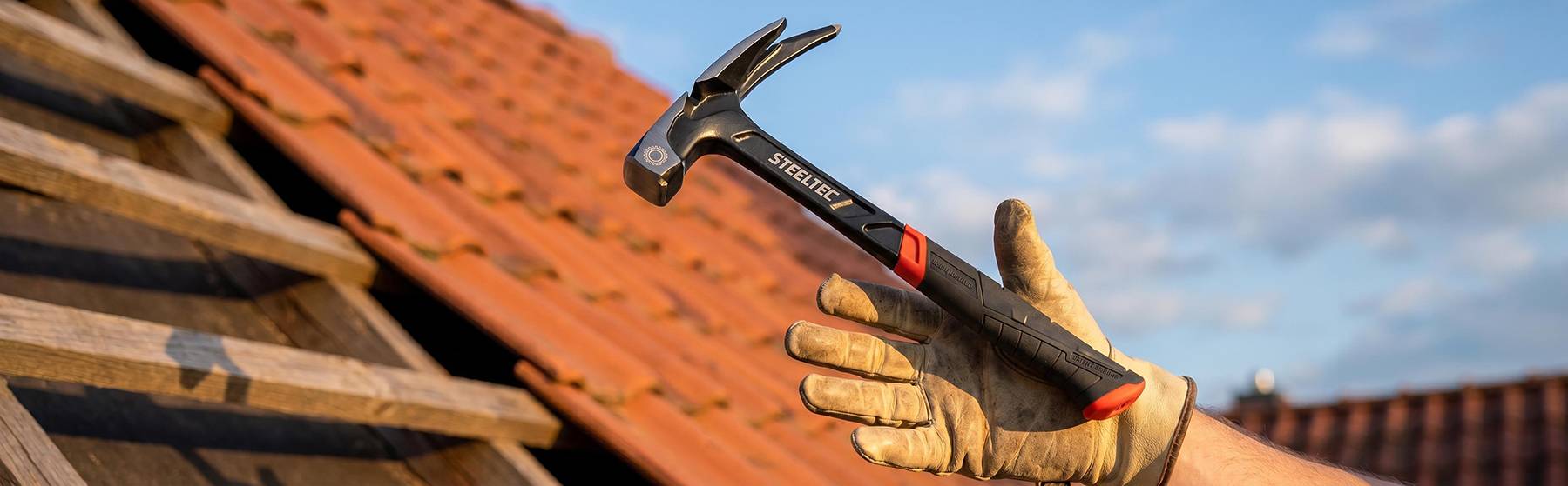 Rukavicí obalená ruka drží tesákovitý kladivo nad červenou střechu z tašek, což naznačuje opravu či stavební práce na střeše.