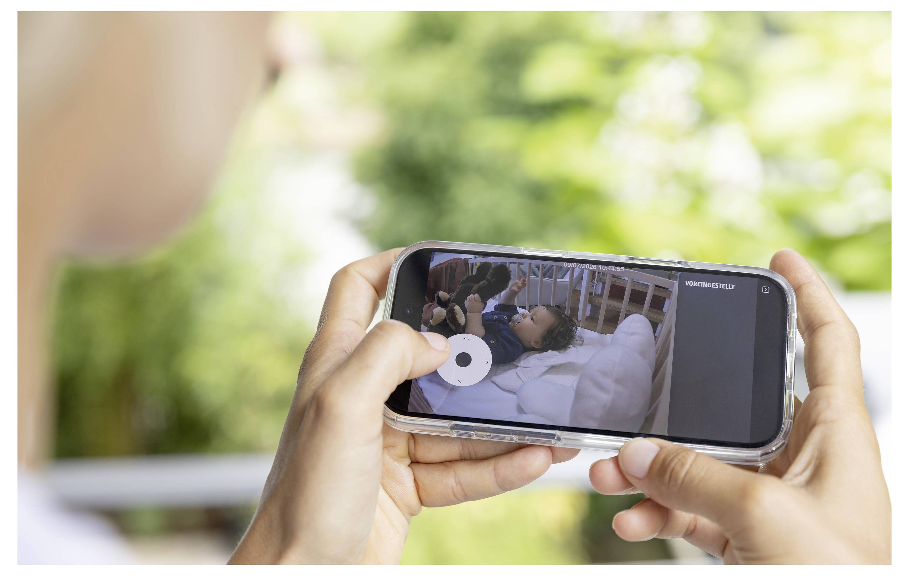 Osoba držící smartphone, který zobrazuje video člověka ležícího v nemniční posteli, sledováno venku před rozostřeným zeleným pozadím.