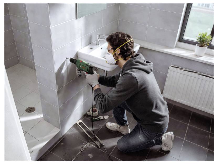 Muž v masce vrtá do stěny v koupelně elektrickým nástrojem během domácí renovace.