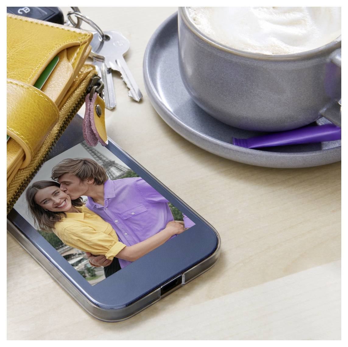 Smartphone na stole zobrazuje pár, který si obejímá na obrazovce, vedle něj je šálek kávy, klíče a žlutá peněženka.