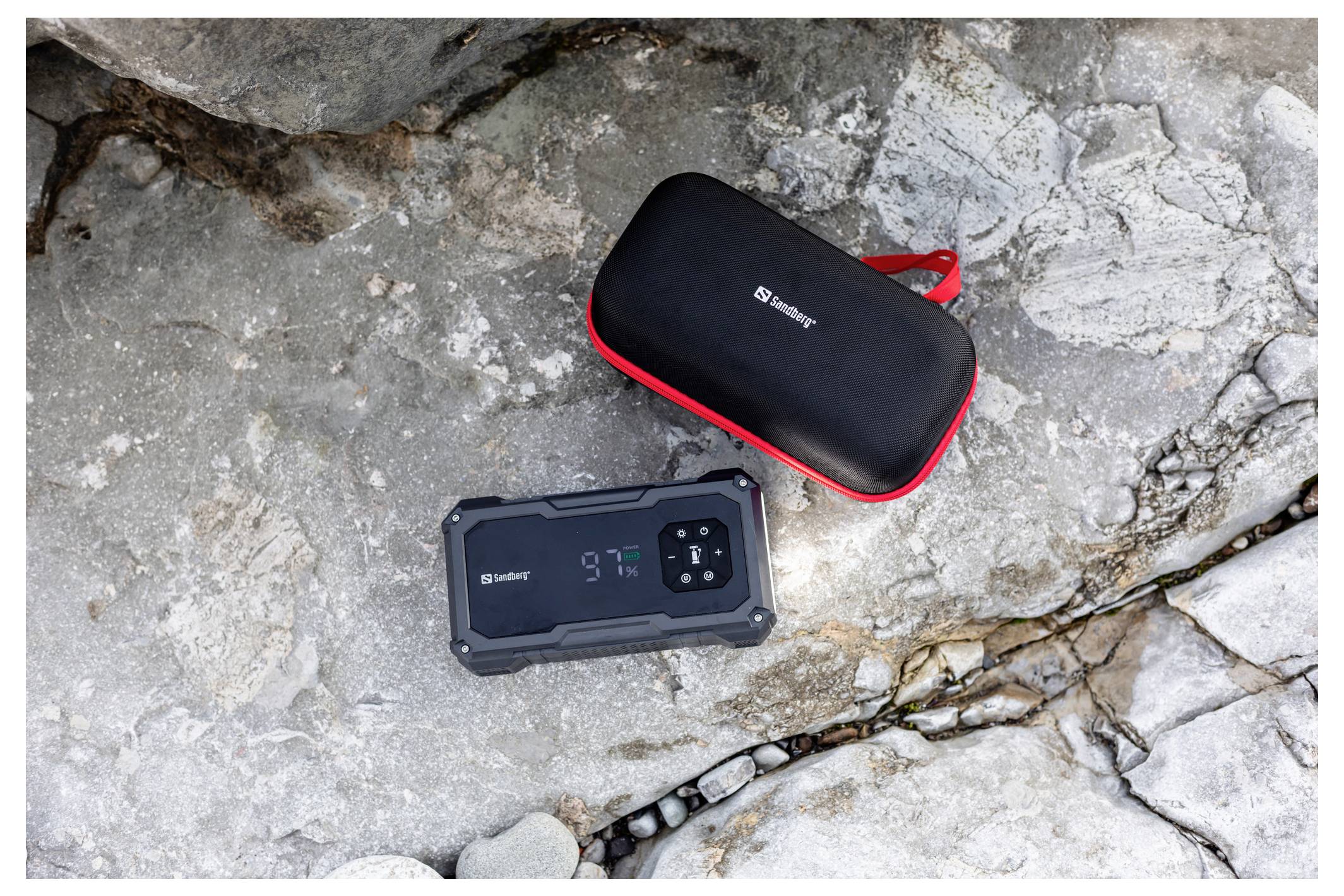 Odolný přenosný Bluetooth reproduktor a odpovídající brašna se zipem ležící na kamenitém povrchu, přičemž displej reproduktoru zobrazuje stav baterie.