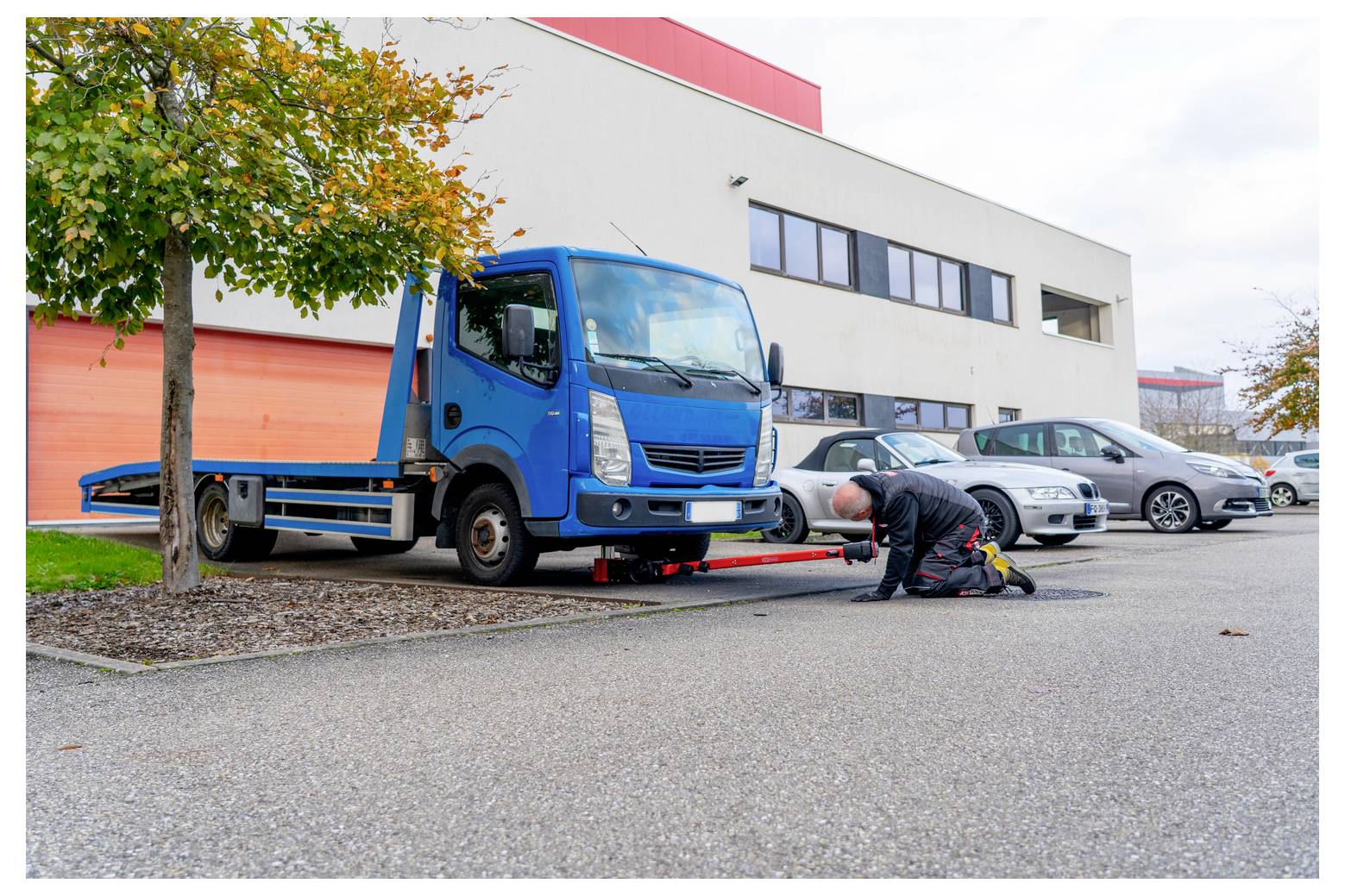 Mechanik klečí pod modrou odtahovací vozidlem zaparkovaným vedle průmyslové budovy a kontroluje nebo opravuje vozidlo.