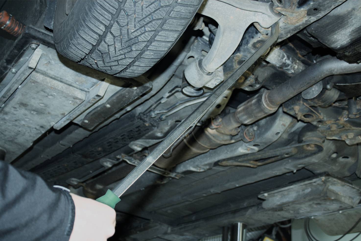 Mechanik ukazuje šroubovák na spodní část automobilu blízko výfuku a komponentů podvozku na zvedáku.