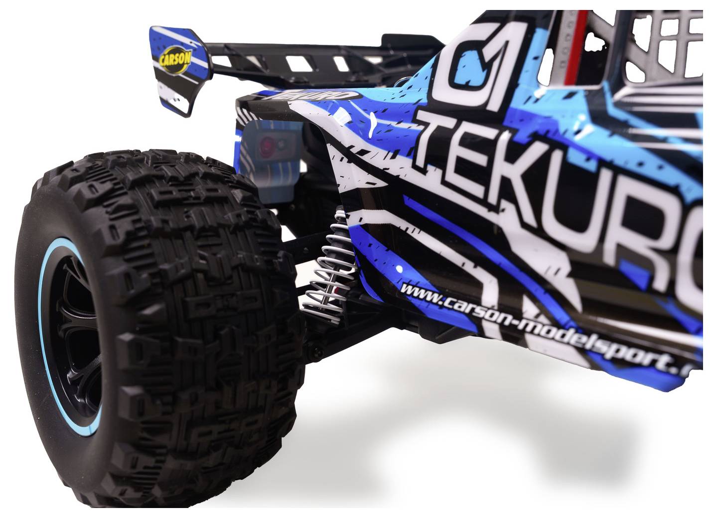 Detailní záběr RC terénního buggyho zobrazující velkou hrubou zadní pneumatiku, odpružení se spirálou a modro-černou karosérií se značkou "TEKNO".