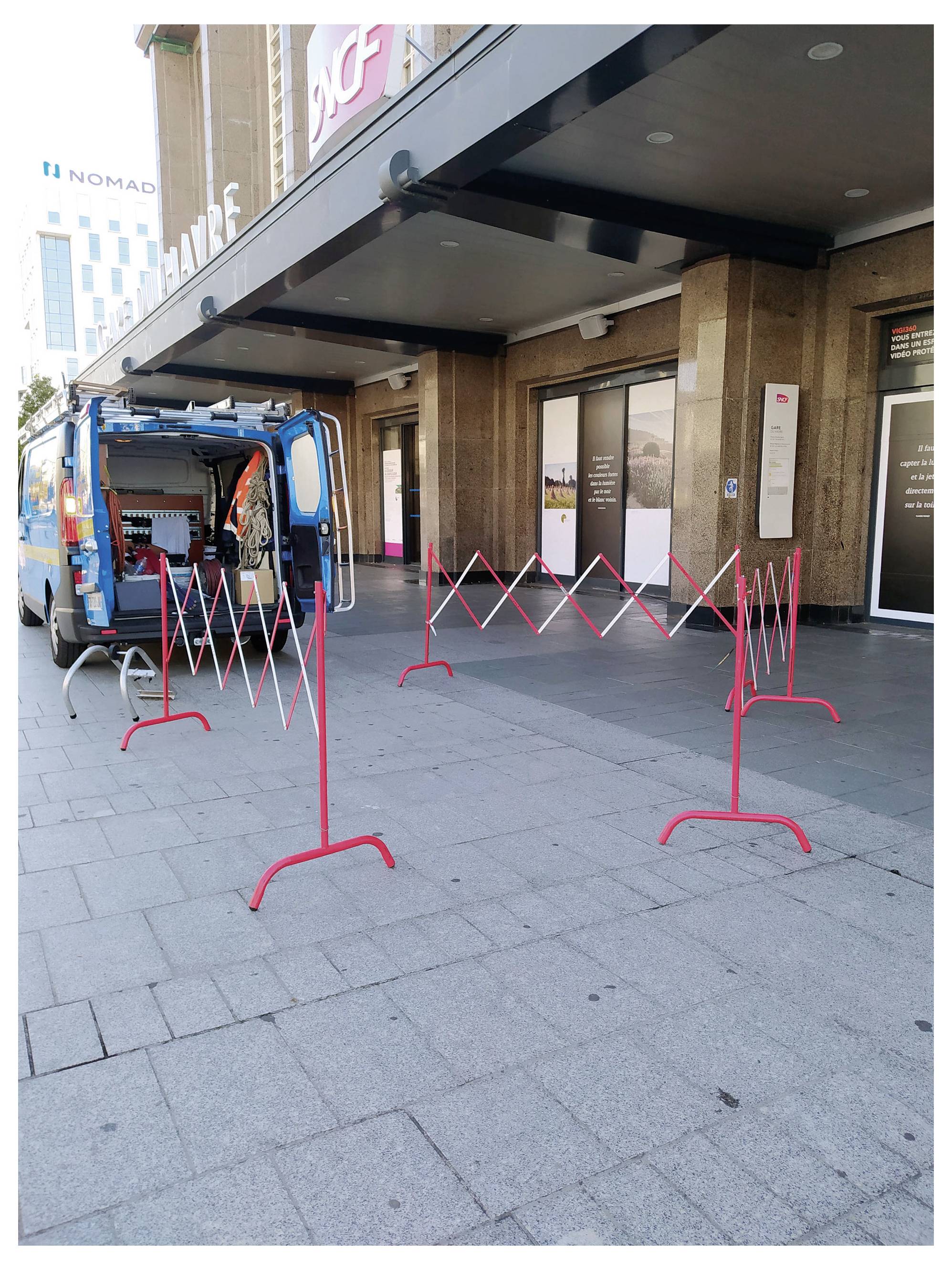 Modrý servisní vůz zaparkovaný vedle vstupu do budovy s červenými zábranami pro kontrolu davu blokujícími přístup na chodníku.