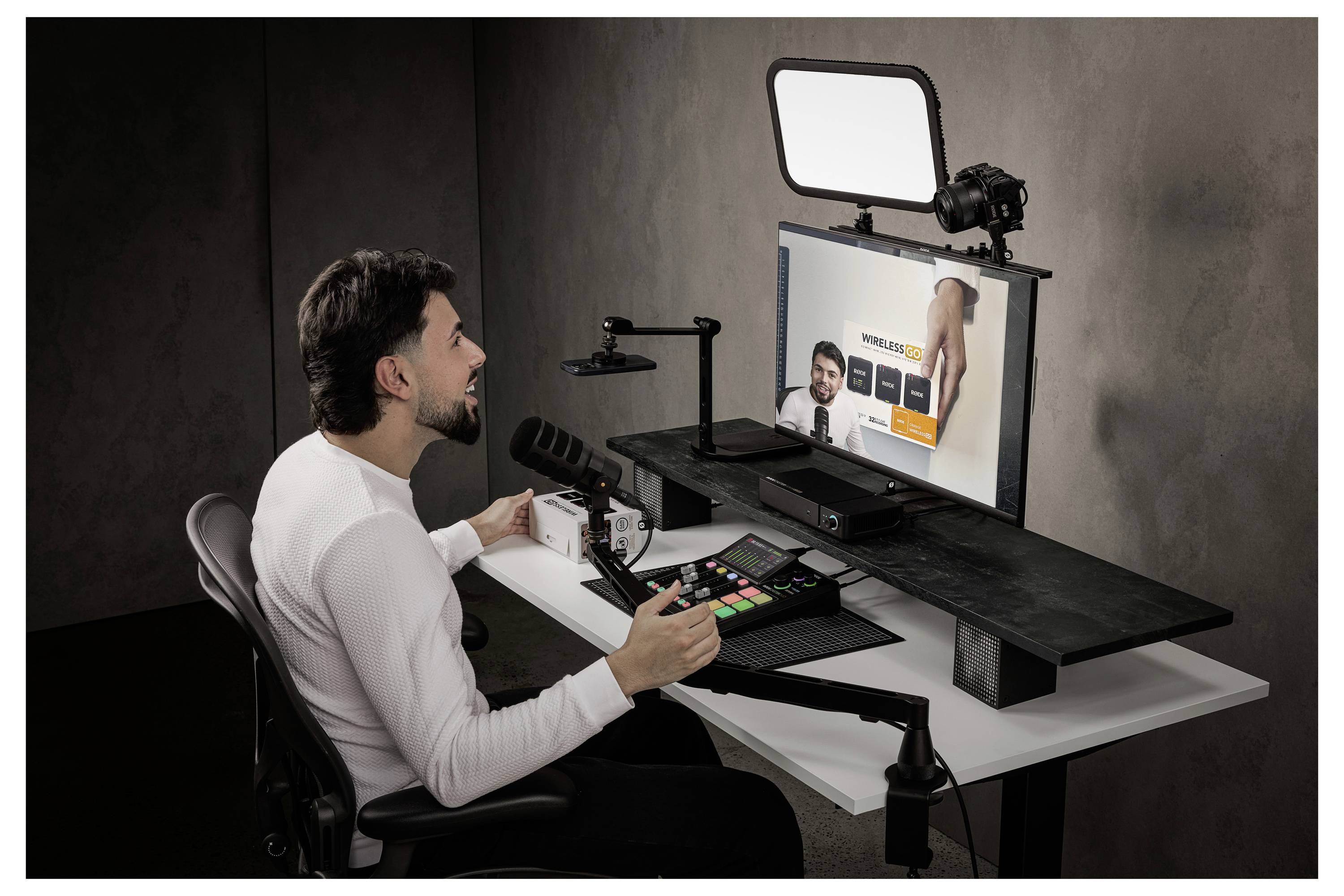 Muž vysílá v přímém přenosu u stolu s kamerou, osvětlením, mikrofonem a monitorem zobrazujícím videohovor nebo online prezentaci.