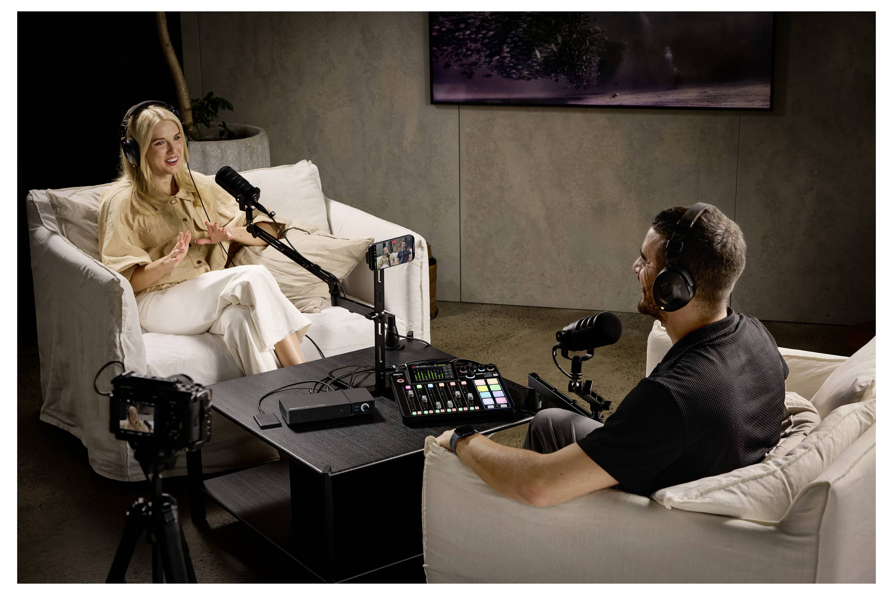 Dva lidé nahrávají podcastový rozhovor v obývacím pokoji, sedí na bílých pohovkách s mikrofony, kamerou a audio mixerem mezi nimi.
