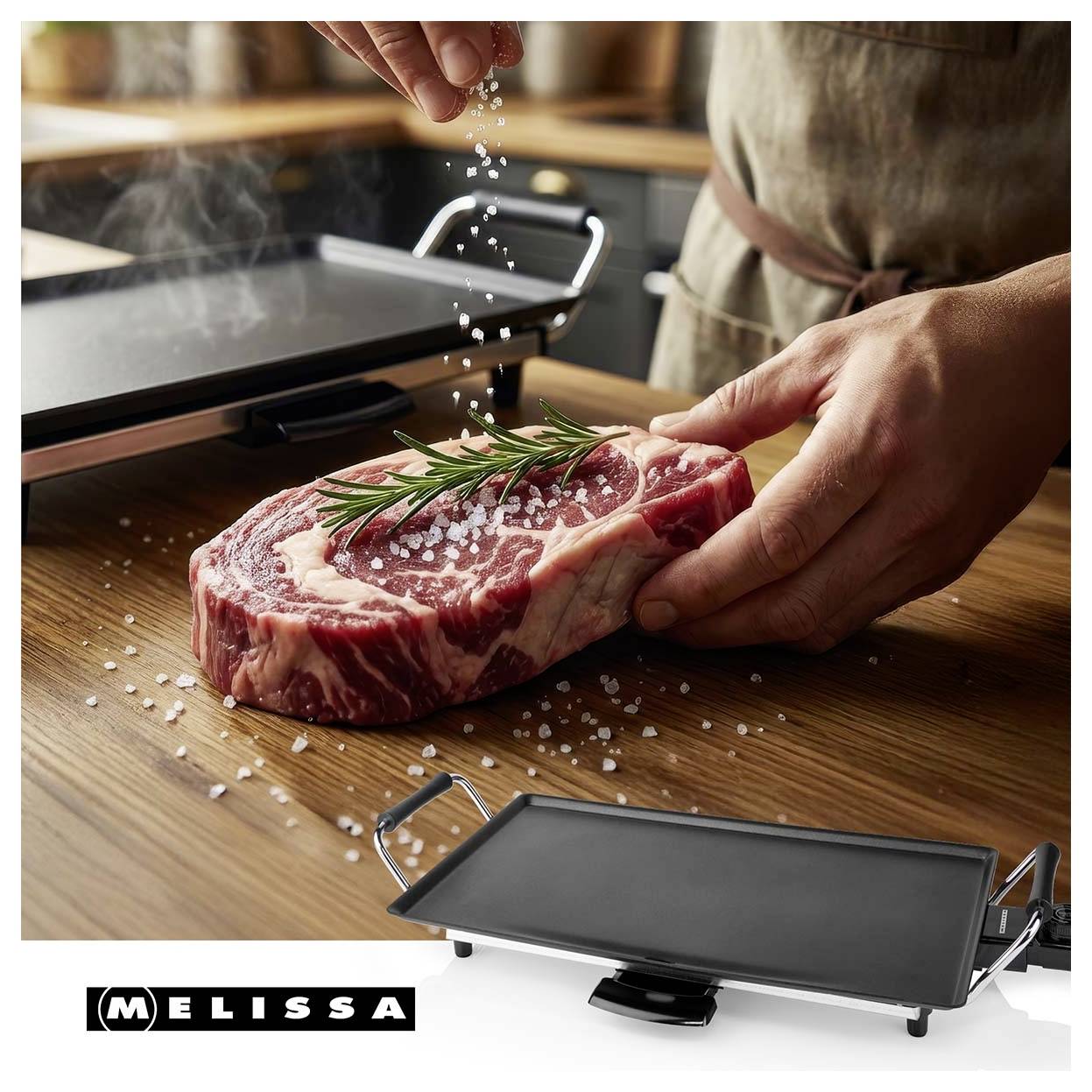 Osoba osolující surový steak rozmarýnem na dřevěném pultu vedle elektrického grilovacího stolu MELISSA.