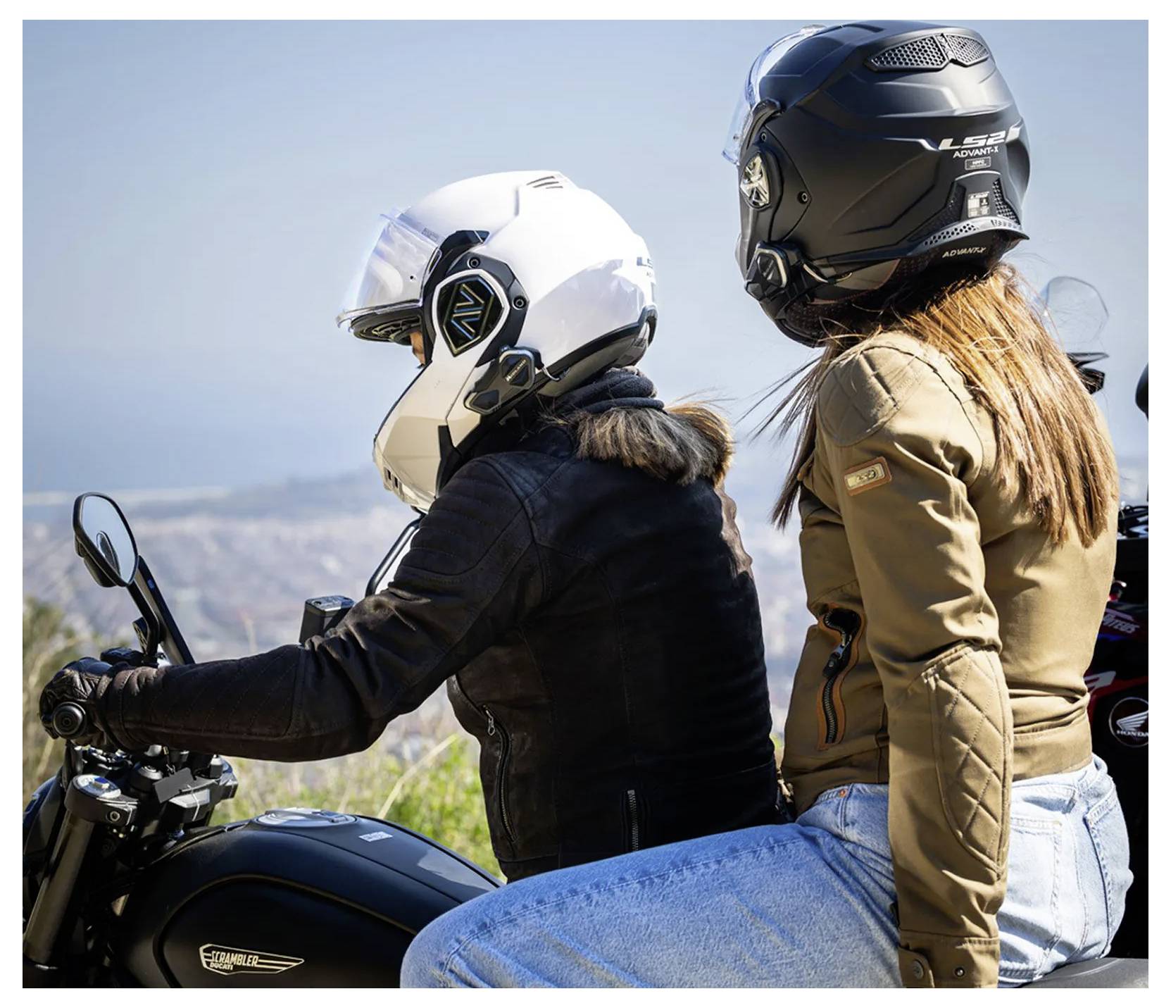 Dvě ženy v přilbě sedí na motocyklu, jedna řídí a druhá sedí na zadním sedadle, v pozadí jsou hory.