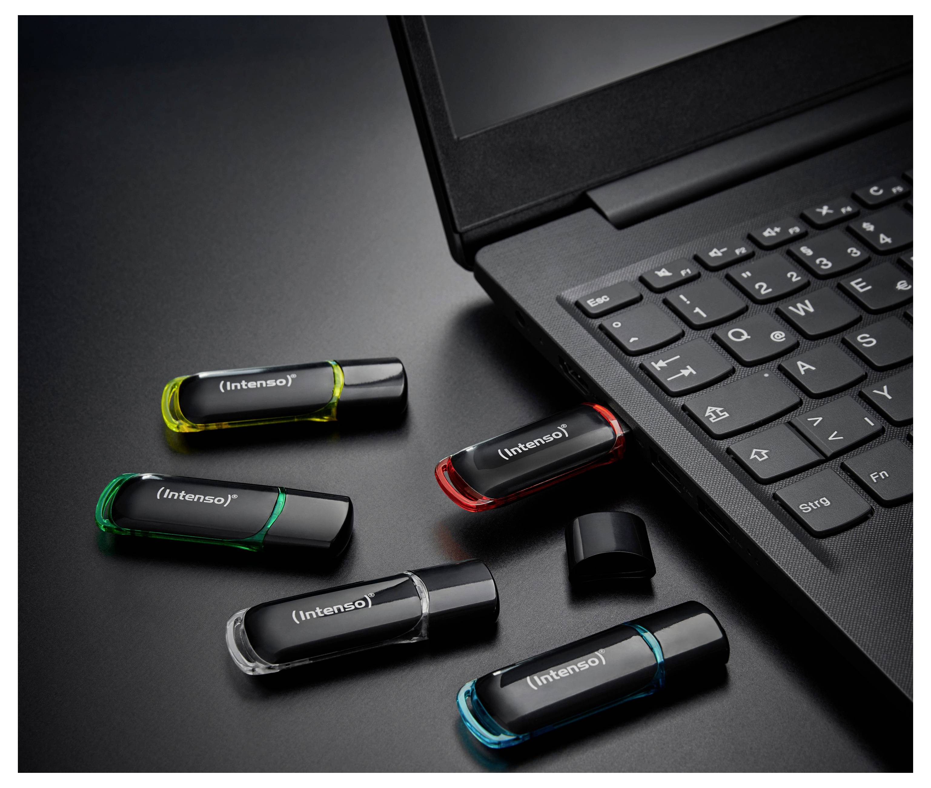 Pět USB flash disků Intenso v různých barvách vedle klávesnice notebooku na tmavém povrchu.